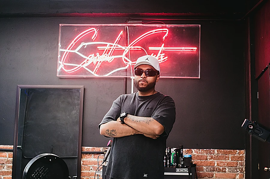 Barbero de Cartel Cuts con gorra blanca y lentes oscuros, de brazos cruzados frente al neón rojo con el nombre de la barbería y una pared de ladrillo