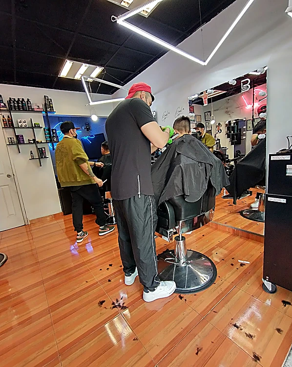 Barbero de gorra roja cortando el cabello a un cliente de espaldas, en la zona de piso de madera con luces blancas en el techo