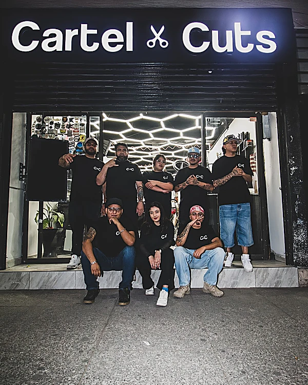 El equipo de Cartel Cuts con playeras negras C×C, de noche en el escalón de la fachada bajo el letrero encendido