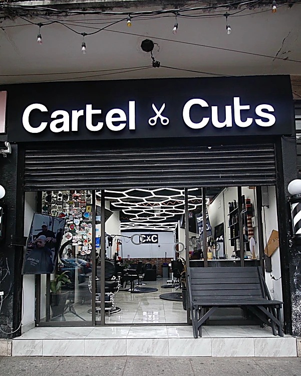 Fachada de Cartel Cuts de día: letrero negro con la tijera entre las dos palabras, postes de barbero a los lados y la puerta abierta al techo de panales de luz