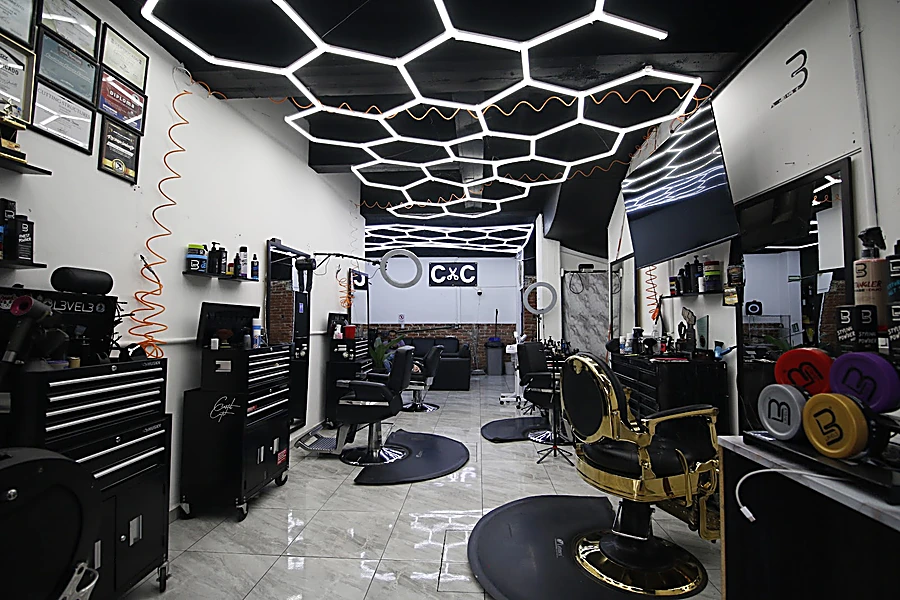 Interior de Cartel Cuts: techo de panales de luz blanca, sillones negros, cajoneras de herramientas, una silla de barbero dorada y piso de mármol
