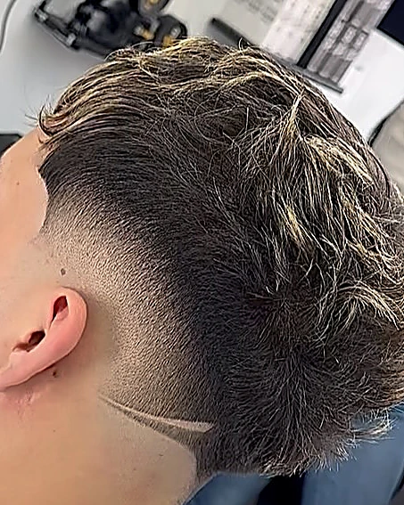 Fade alto con una línea rasurada en diagonal detrás de la oreja y el cabello de arriba texturizado con puntas claras